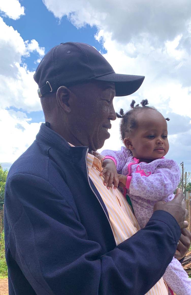 Andrew in a cap holding a baby in a purple outfit under a bright sky