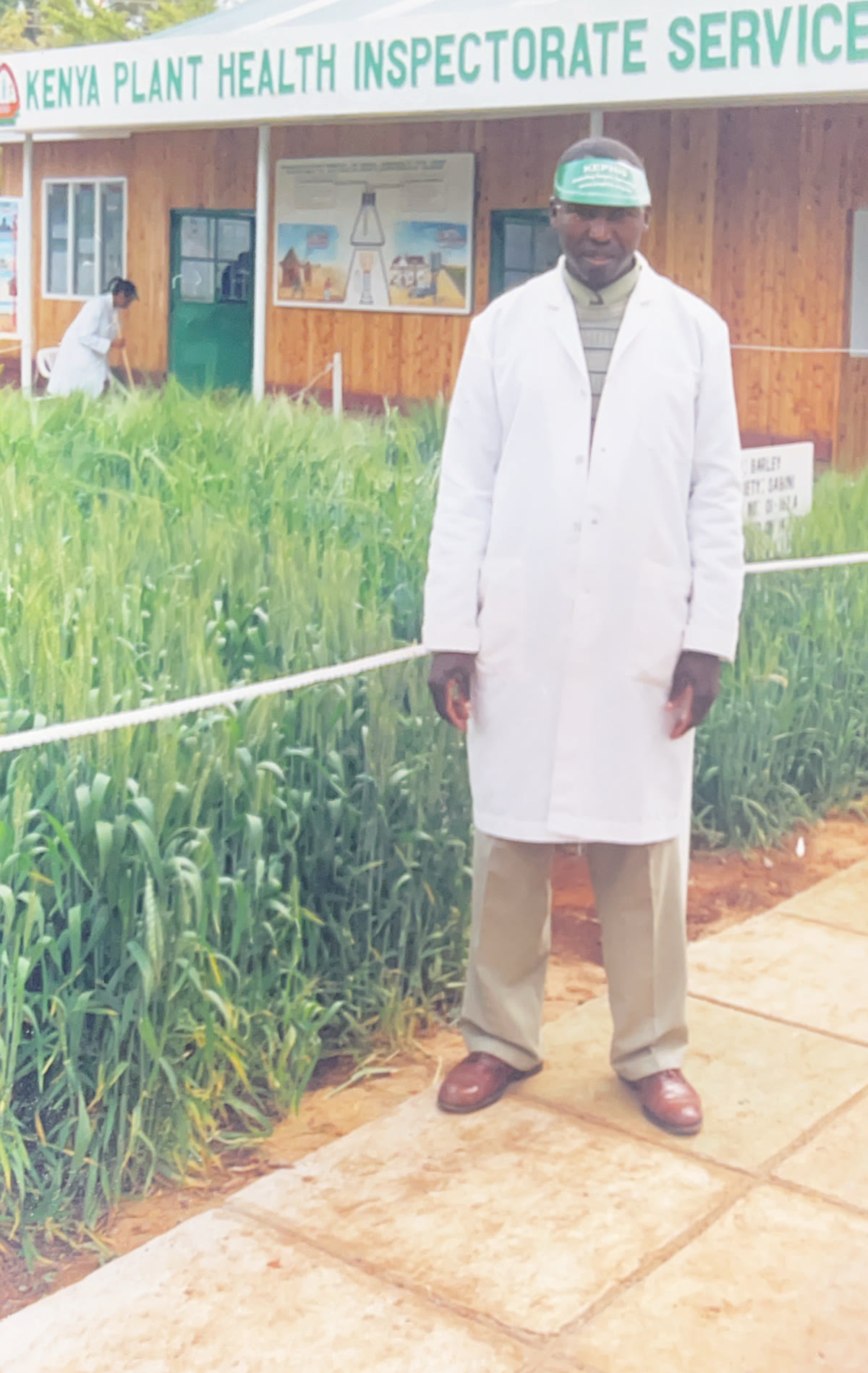 Andrew wearing a white lab coat beside a crop display at the Kenya Plant Health Inspectorate Service.