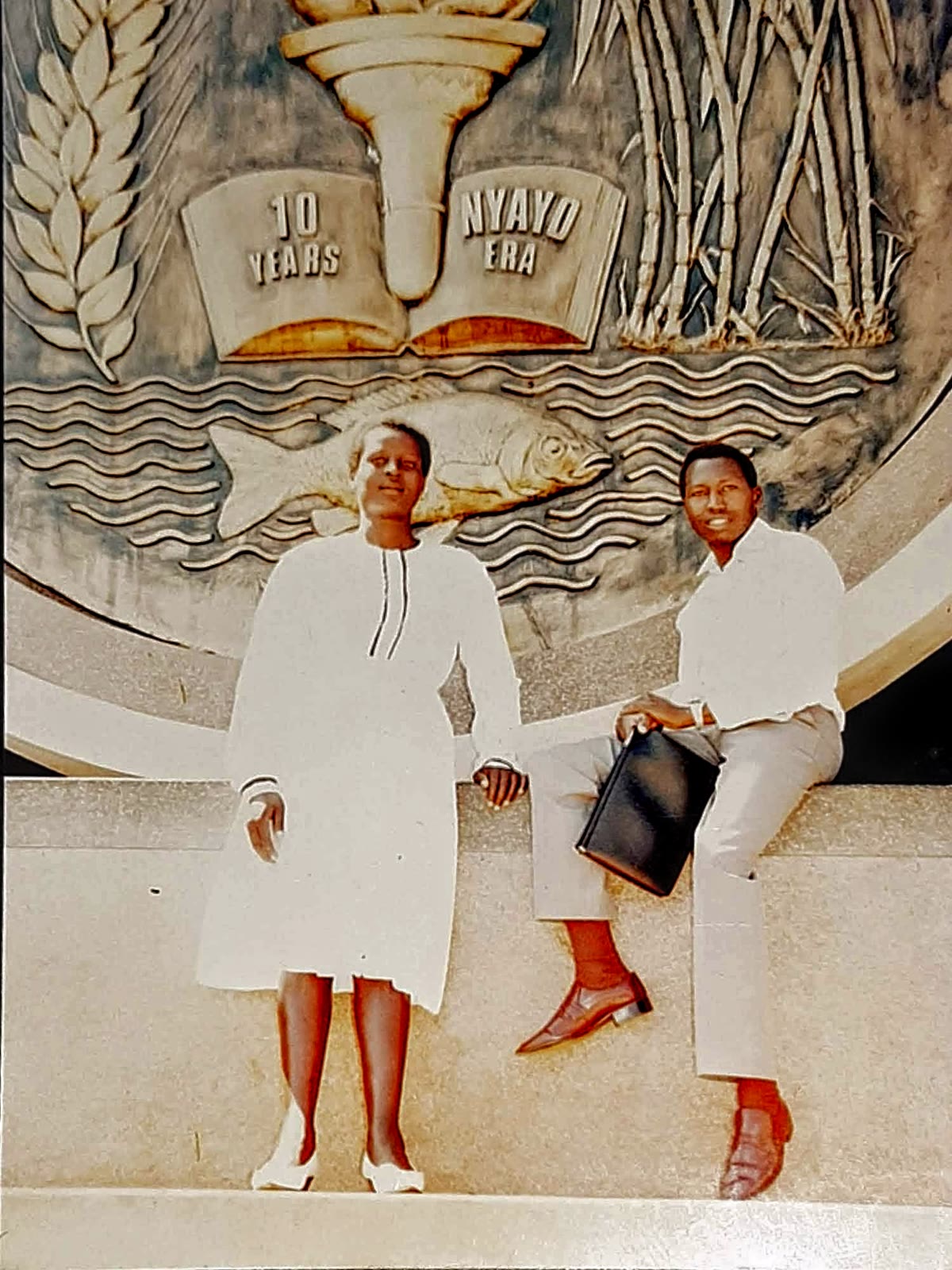 Two people posing in front of a monument marked 10 Years Nyayo Era.
