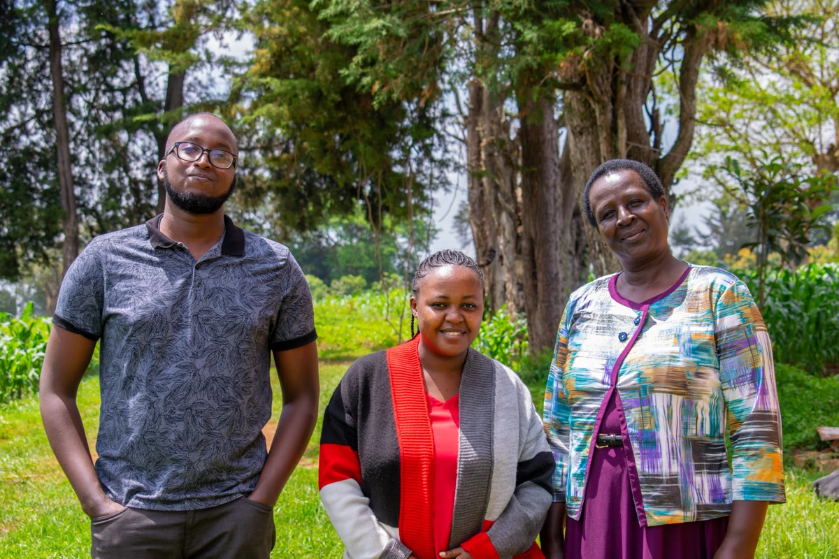 Three family members standing together outdoors beneath trees.